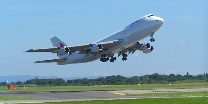 A large white Boeing 747 jumbo jet taking off from an airport runway against a clear blue sky, with its landing gear still extended.