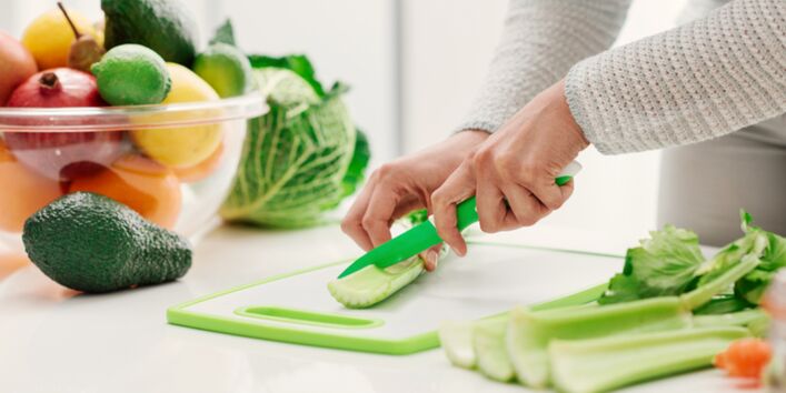 Foto. Ein Mensch schneidet grünen Sellerie auf einem Schneide-Brett. Daneben steht eine große Schüssel mit buntem Obst und Gemüse, wie Avocados, Zitronen und Äpfeln.