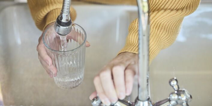 High-angle, close-up shot of hands wearing a yellow sweater, holding a clear ridged glass and filling it with fresh water from a modern chrome kitchen faucet.