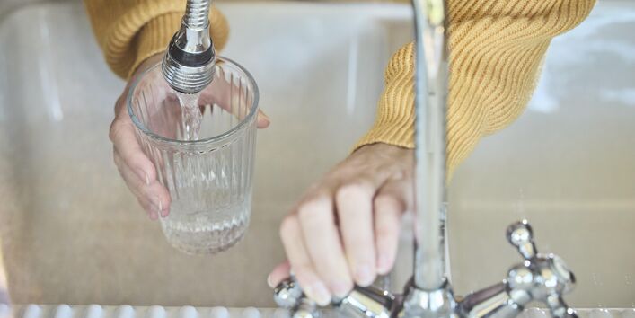 Foto. Ein Mensch in einem gelben Pullover steht an einer Spüle. Er hält ein Glas unter einen Wasser-Hahn. Mit der anderen Hand dreht er das Wasser auf.