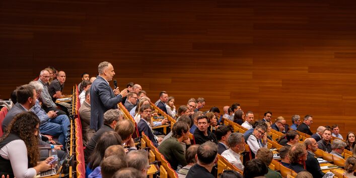 Eine Person aus dem Publikum beteiligt sich mit dem Saalmikrophon in der Hand an der Diskussion im UBA-Hörsaal. Mehrere Menschen blicken interessiert in die Richtung der Person.