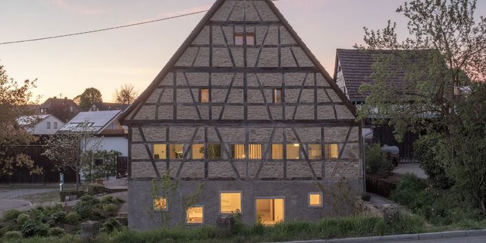 : Foto der Giebelwand eines viergeschossigen Fachwerkhauses mit Gefachen aus Feldstein und Satteldach. In der dörflichen Szene während der Dämmerung sind die Fenster mit warmem Licht erleuchtet.