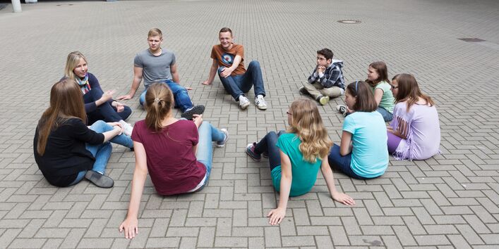 Auf dem Bild sind Jugendliche zusehen, die in einem Kreis auf dem Schulhof sitzen.