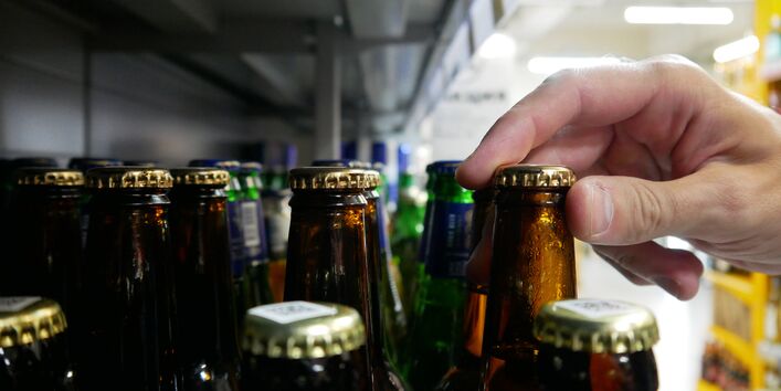 Eine Hand greift in einem Supermarktregal nach dem Kronkorken einer braunen Glasflasche, die zwischen anderen braunen und grünen Flaschen steht.
