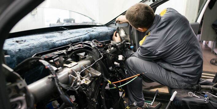Ein Mechaniker in grauer Arbeitskleidung sitzt auf dem Beifahrersitz eines Autos und arbeitet am zerlegten Armaturenbrett. Das Innere des Fahrzeugs ist offen gelegt, wobei zahlreiche Kabel, Steckverbindungen und die Metallstruktur der Instrumententafel sichtbar sind.