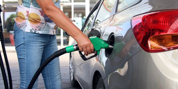  Eine Person in Jeans und einem T-Shirt mit Blumenmuster hält eine grüne Zapfpistole in den Tank eines silberfarbenen Autos.