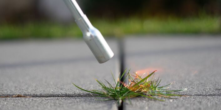 Ein Abflammgerät aus Metall brennt Gras in einer Fuge ab.