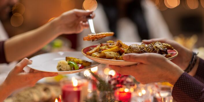 Eine Person serviert einer anderen Person gegartes Gemüse auf den Teller. Es herrscht ein weihnachtliches Ambiente.