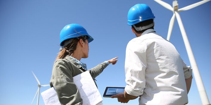 Das Bild zeigt im Vordergrund zwei Ingenieure, welche sich über Pläne beugen. Im Hintergrund sind Windenergieanlagen zu sehen. 