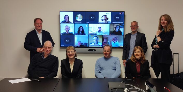 In a small meeting room, 7 people sit or stand in front of a screen. On the screen appear the faces of people in the meeting call.
