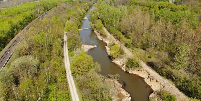 Foto: Gezielt eingebrachte Strömungslenker in Form von quer zur Fließrichtung verlaufenden Buhnen aus großen Steinen. Dichte Vegetation wächst entlang der Ufer, die einen befestigten Weg und in etwas größerer Distanz Eisenbahnschienen vom Gewässer trennt.