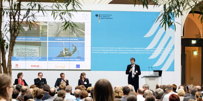 Ein Mann steht und spricht auf dem Podium, drei weitere Personen sitzen 