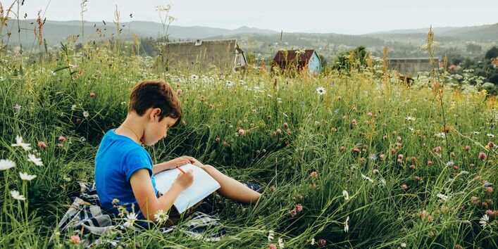 Ein Junge sitzt in einer Blumenwiese mit Blick auf eine grüne hügelige Landschaft.