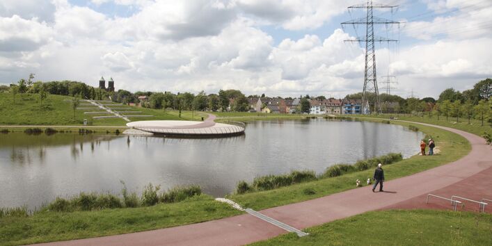 Grünfläche mit See, im Hintergrund ist ein Wohngebiet zu erkennen, Menschen spazieren am See entlang