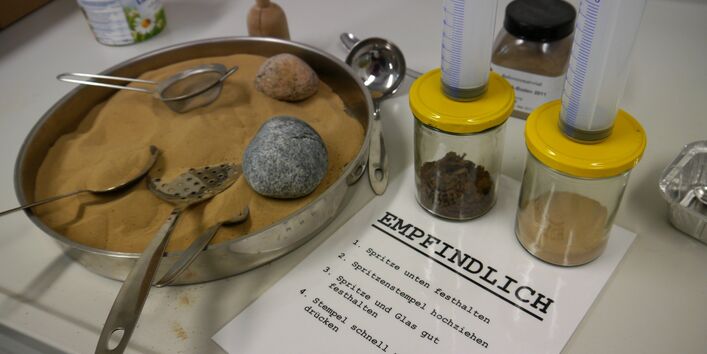 Auf einem Tisch stehen zwei Gläser mit Boden, in die mit jeweils einer großen Spritze Luft gepustet werden kann. Daneben steht feiner Sand, der durch ein Sieb rinnt.