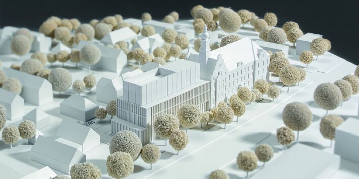 ein Architekturmodell zeigt ein historisches viergeschossiges Gebäude mit Giebel und Türmchen und einen ebenso hohen modernen Anbau inmitten einer Stadt mit Straßen, Bäumen und niedrigeren Häusern