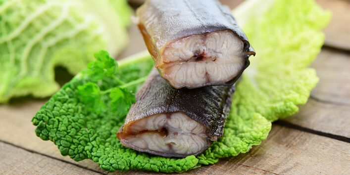 Zwei Stücken Aal auf einem Wirsingkohlblatt liegen auf einem Holztisch, im Hintergrund Messer und Gabel