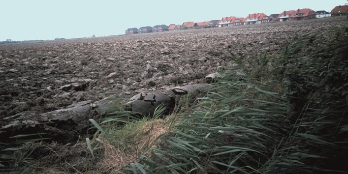 ein kahler, umgepflügter Acker, im Hintergrund ein Neubaugebiet