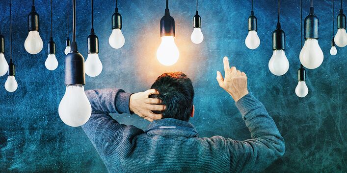 Ein Mann steht mit dem Rücken zum Betrachtenden. Kratzt sich am Kopf und zeigt mit der rechten Hand in die Luft. Über ihm hängen viele Glühbirnen, von denen eineüber seinem Kopf leuchtet.