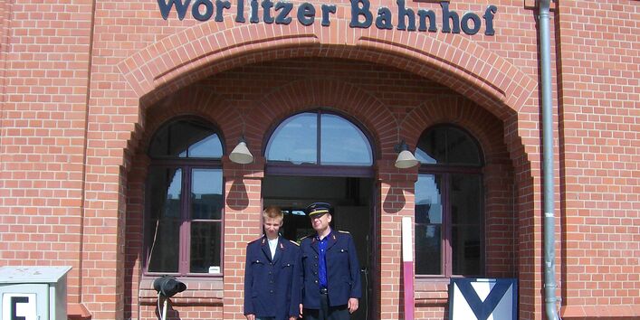 zwei Männer mit alten Bahnmitarbeiter-Uniformen vor dem Eingang des Wörlitzer Bahnhofs