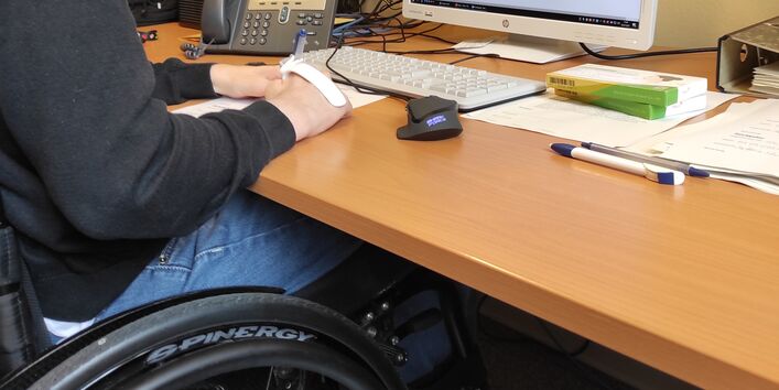 ein junger Mann sitzt im Rollstuhl im Büro an einem Schreibtisch 