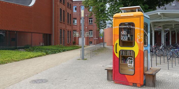 eine bunt angemalte Telefonzelle ist innen zu einer Miniblibliothek ausgebaut, auf dem Dach ein Solarmodul, außen eine Holzbank