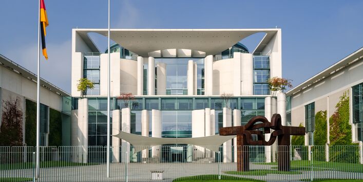 modernes weißes Gebäude mit Flachdach und großzügigem Eingangsbereich mit verglaster Fassade, davor eine deutsche Flagge und eine moderne Gartengestaltung mit amorphen kleinen Rasenflächen und modernen Skulpturen