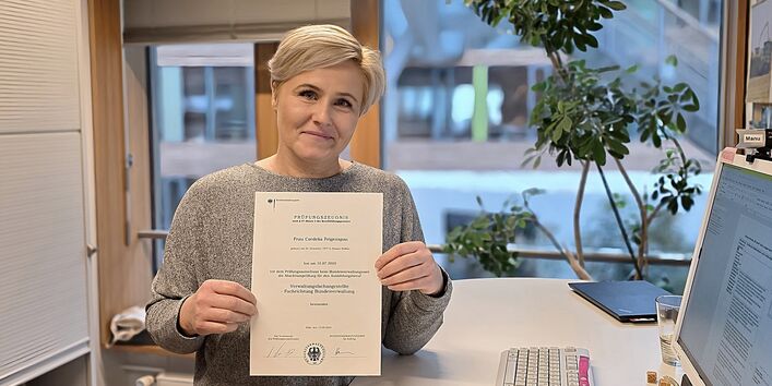 Cordelia Feigenspan hält in ihrem Büro neben ihrem Schreibtisch stehend ihr Prüfungszeugnis in die Kamera