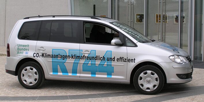 ein silbergrauer VW Touran steht vor dem UBA Dessau-Roßlau. Er trägt die Beschriftung "CO2-Klimaanlagen - klimafreundlich und effizient, R744" und das Logo des Umweltbundesamtes