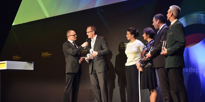 auf einer Bühne: ein Mann im Anzug hält eine Blauer Engel-Trophäe in der Hand und wird mit einem Mikrofon befragt, daneben in einer Reihe weitere Personen