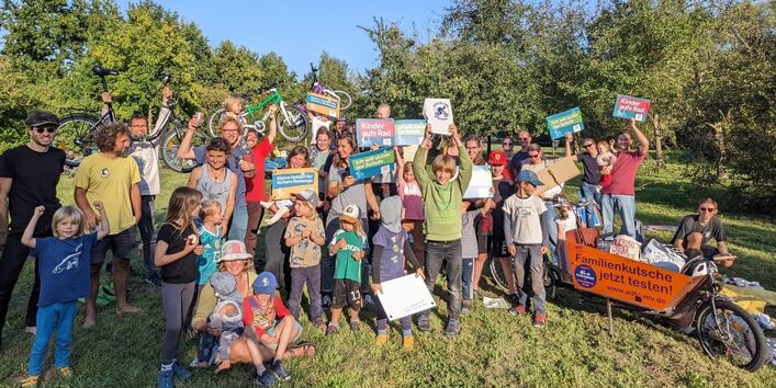 Gruppe Erwachsener und Kinder mit Plakaten und einem Lastenrad auf einer großen Rasenfläche mit Bäumen