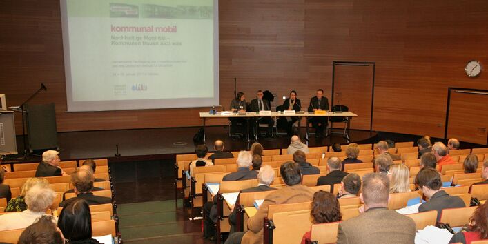Veranstaltung im Hörsaal des UBA-Dessau: auf dem Podium sitzen 2 Männer und 2 Frauen, im Zuschauerraum sitzen Leute mit Tagungsmappen, auf eine Leinwand wird projiziert "Kommunal mobil, Nachhaltige Mobilität - Kommunen trauen sich was"