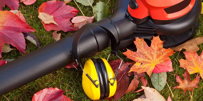 Auf einer Rasenfläche liegen ein Laubbläser, ein Gehörschutz-Kopfhörer und bunte Blätter. 