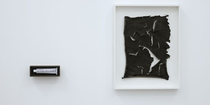 an einer weißen Wand hängt eingerahmt ein kohlrabenschwarzes, zerrissenes Blatt Papier oder Haut, daneben hängt, schwarz eingerahmt, eine Tube mit der Aufschrift "Neckartorschwarz" 
