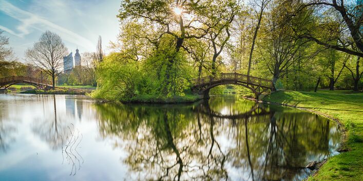 in einem Park spiegeln sich Bäume und eine Brücke in einem Teich, im Hintergrund Gebäude einer Großstadt