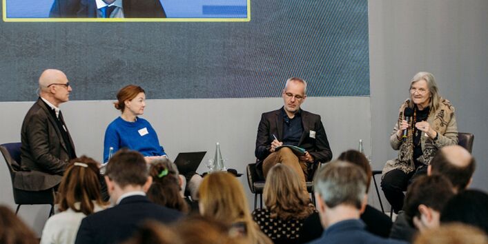 Vier Leute sitzen auf Stühlen auf einer Bühne und reden miteinander, man sieht die Rücken der im Publikum sitzenden Teilnehmer*innen.