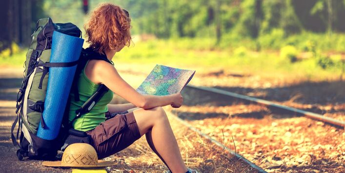 eine junge, sportlich gekleidete Frau mit Rucksack, Strohhut und Isomatte wartet auf der Bahnsteigkante in der Sonne sitzend auf einen Zug und schaut in eine Landkarte