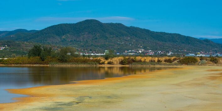 zum Teil wassergefülltes Rückhaltebecken mit naturnahem Ufer, im Hintergrund bewaldete Berge