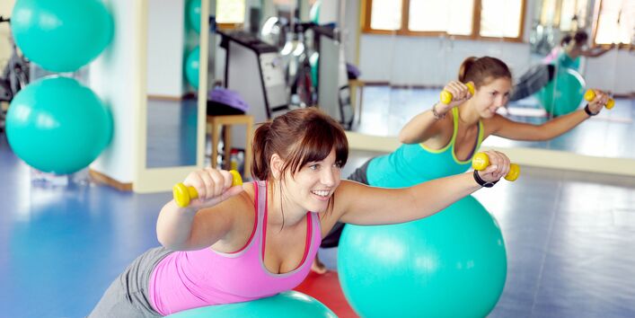 zwei Frauen in einem Sportraum trainieren mit Gymnastikbällen und Hanteln auf einer Matte