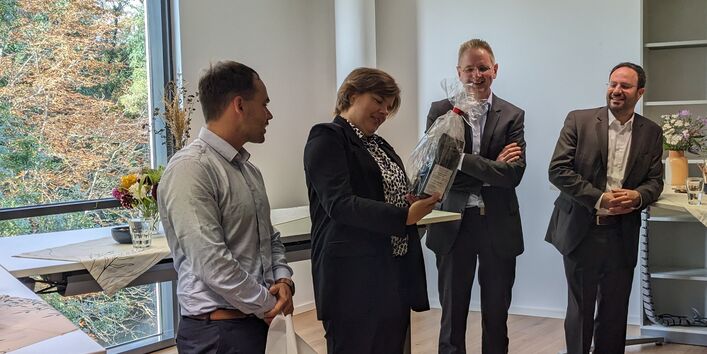 drei Männer und eine Frau mit Geschenk in einem hellen Veranstaltungsraum