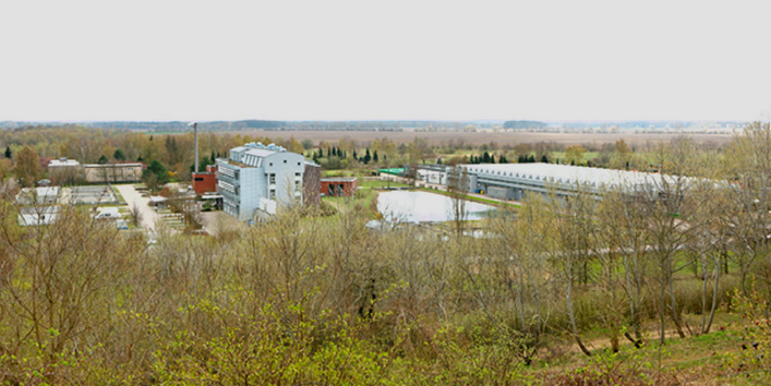 Foto: In Mitten von Bäumen und Feldern ist ein Ensemble aus Gebäuden, einer Halle und Wasserbecken zu sehen.