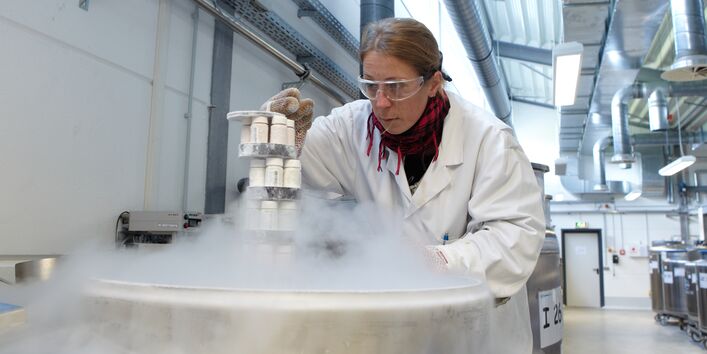 eine Frau in einem weißen Kittel, mit Schal, Handschuhen und Schutzbrille holt Proben aus einem dampfenden Metallbehälter, der in einer Halle steht