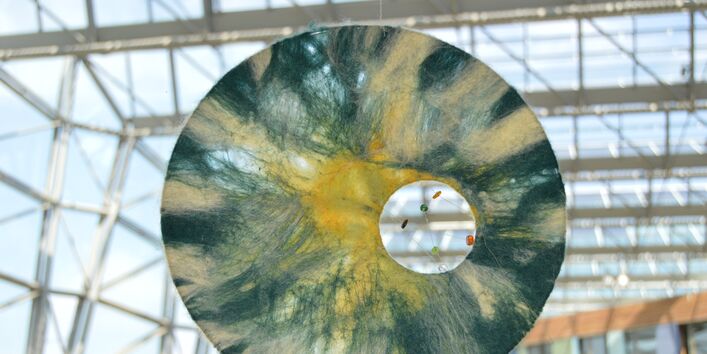 an einer Glasfront des UBA Dessau hängt eine halbtransparente Scheibe, die an Sonne, Mond, Himmel und Wolken erinnert. Ein Guckloch bietet einen Einblick ins Innere des Gebäudes