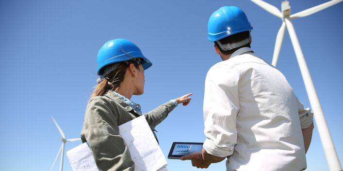 eine Frau und ein Mann mit Bauhelm, Plänen und Tablet schauen sich einen Windpark an