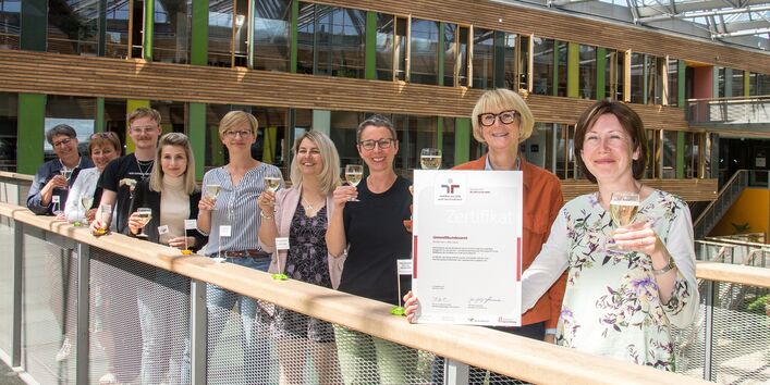 acht Frauen und ein Mann haben sich im UBA Dessau für ein Foto aufgestellt und halten lächelnd Gläser zum Anstoßen und das Zertifikat des Audits Beruf und Familie in die Kamera