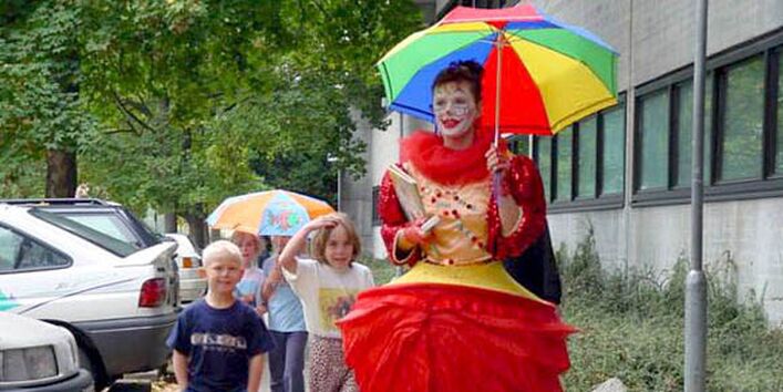 Eine Frau als Clown verkleidet hält einen bunten Regenschirm und läuft mit einigen Kindern auf einem Bürgersteig.