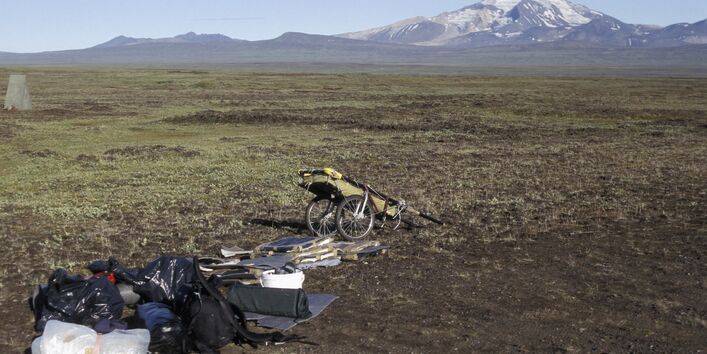 Eine weite Steppenlandschaft, im Hintergrund schneebedeckte Berge. Im Vordergrund der Handwagen der Künstlerin mit ihrem Gepäck.