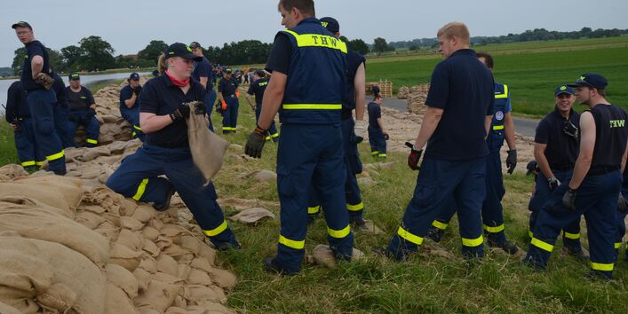 Personen des THW beim Errichten eines Deichs aus Sandsäcken in Flußlandschaft.