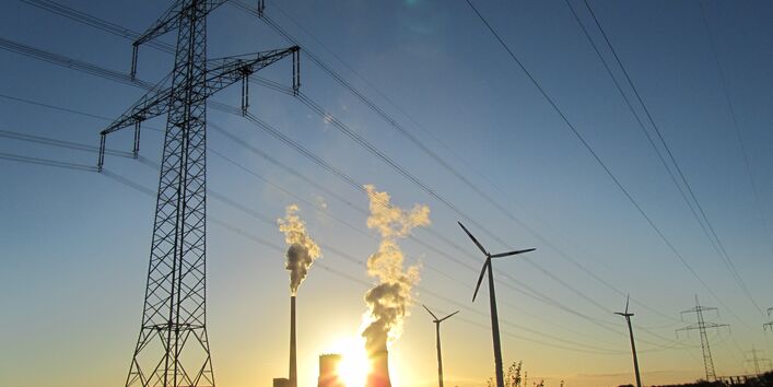 Übertragungsmasten mit Hochspannungsleitungen, Windräder und konventionelles Kraftwerk vor Sonnenuntergang.