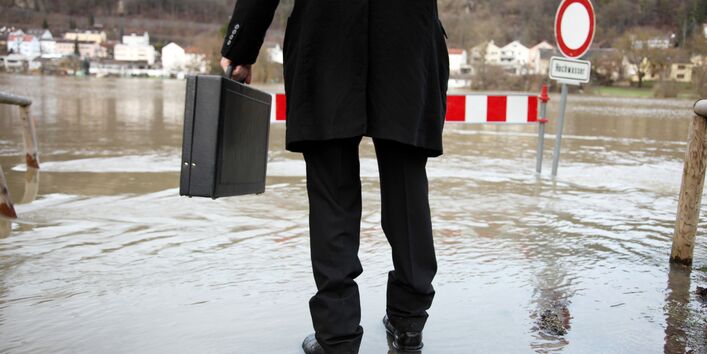 Rückenaufnahme eines Mannes im schwarzen Anzug mit Aktentasche, der auf überfluteter Fläche steht.